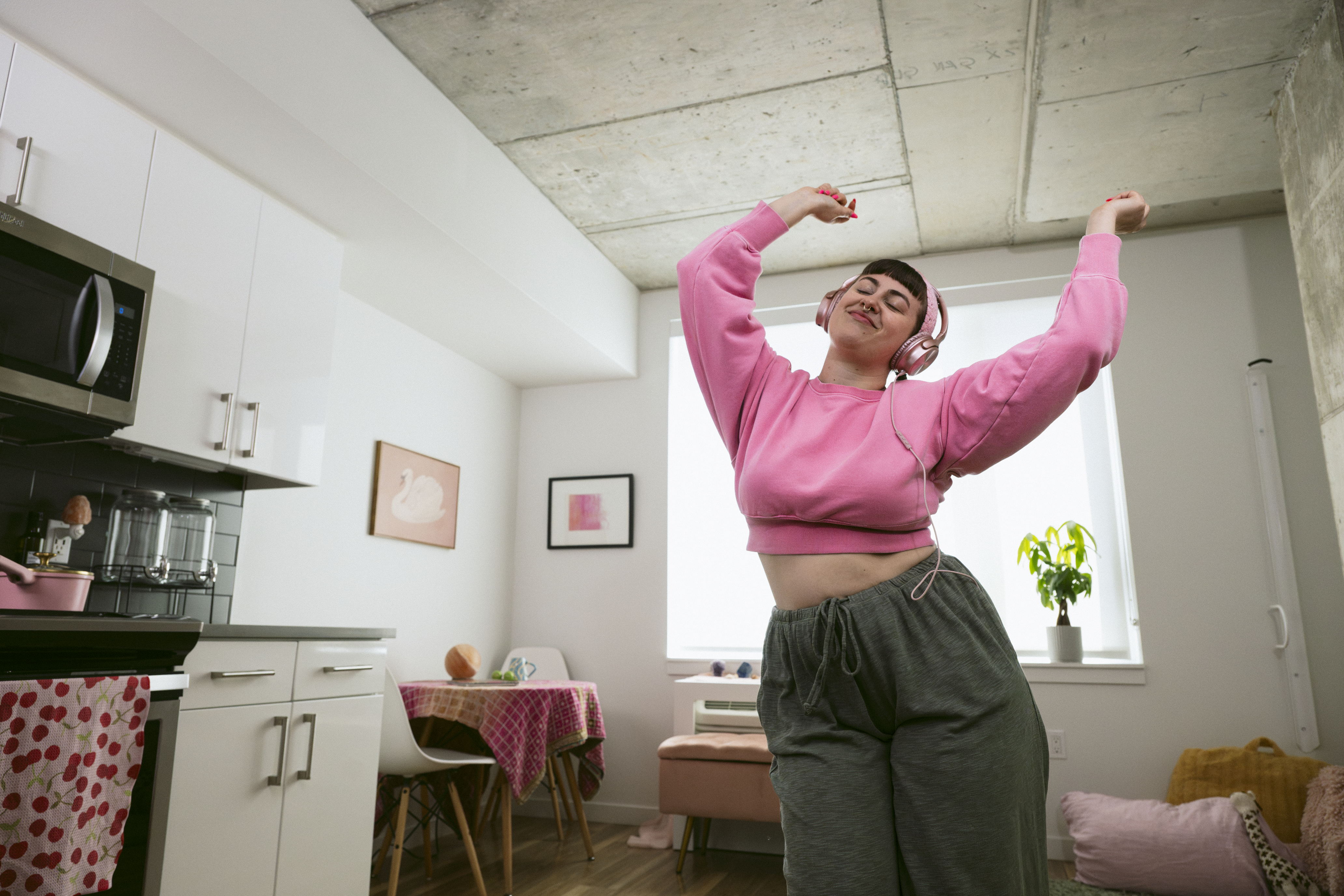 Mujer con sudadera y pantalones rosas bailando en su sala de estar y usando auriculares.